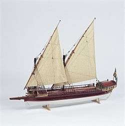 A side view of a model of a small galley with two masts rigged with lateen (triangular) sails. Its outrigger folded up and the oars stowed on the deck. The hull above the waterline is painted red with decorative details in gold and blue. The bow has a raised platform (rambade) armed with 3 small cannons.