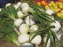 A pile of onions, with green stems protruding from white bulbs. Organes, lemons and limes are visible in the background.