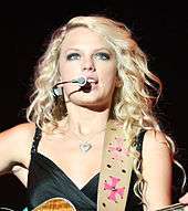 A blonde woman wearing a dark shirt is playing the guitar and singing