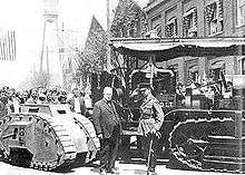 A military man in full dress uniform salutes a suited business man who is doffing his bowler hat in return. Behind the two men are a Holt tractor, which towers above them, and a diminutive one-man tank, which is below chest height. Behind the vehicles, a crowd looks on.