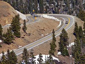 =A sharply curved section of road, viewed from above, in a steeply sloped area with large evergreen trees