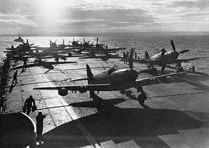 The aft section of an aircraft carrier's flight deck. Several propeller aircraft are parked here, and are being worked on.