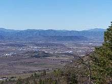 Vegetation in the foreground, with a flat area scattered with buildings farther away. The plateau is far away, with mountains by the horizon.