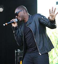 A African-British man, wearing black sunglasses and dark jacket, shirt and trousers, sings into a microphone.