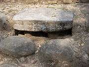 A thick, flat stone set into a wide step, it has a dark hollow underneath with two short stone legs supporting the altar. Two large, smoothly rounded cobbles are set into the step below the altar.