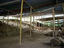 A cleared area of bare soil strewn with rubble, with raised areas to the left and right and roofed with corrugated metal supported by timber poles