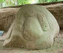 A large boulder set amongst the bare earth of an excavated area, with some vegetation visible in the background. The boulder has been carved into the form of an obese head with prominent puffed-out cheeks and swollen lips. The ears have large ear-spools hanging from them and the eyes are closed.