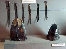 Six long, thin blades of dark volcanic glass, suspended vertically in a display cabinet. In front of them are two angular pieces of dark, shiny stone.