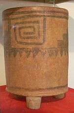 A tall cylindrical ceramic vessel with three stubby legs. The base colour is orange with a darker pattern consisting of a square spiral.