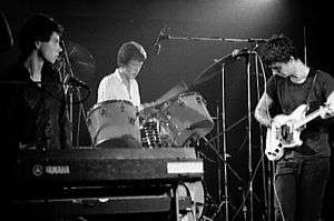 Three men are situated on a stage, each with his own respective instrument: a keyboard, a drum set and a guitar.