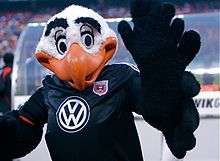 A black and white costumed bald eagle mascot with exaggerated features and an orange beak raising his wings. He wears a black soccer jersey with a white Volkswagen logo and the team's shield on it.