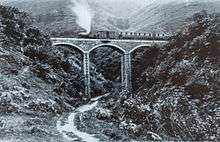 A stone viaduct with three arches spans a steep sided ravine. A steam train stands on the viaduct, with an engine, a van and three coaches.