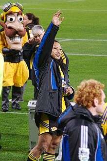 A Polynesian man wearing a yellow rugby jersey and a black jacket with his arm raised