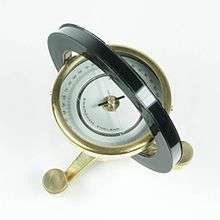 Photograph. The most prominent feature is a horizontal circular compass case that is seen from above. The compass is centered inside of a black ring with a square cross-section. The compass and ring are supported on a brass tripod that has leveling screws as its feet.