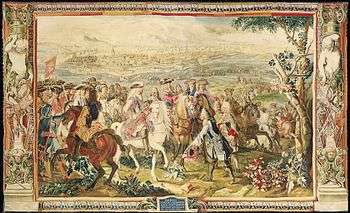 Tapestry shows a crowd of men mounted on horseback. They are dressed in early 18th century style with three-cornered hats and curly shoulder-length wigs. The man in the center rides a white horse, holds a sword and wears a metal cuirass. In front of him, a man on foot has his hat off and slightly bows. A walled fortress city is in the background.