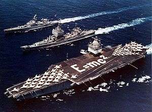 Three ships sale next to each other, including an aircraft carrier which has jets arranged as an arrow and the phrase E=MC2 written with sailors on deck