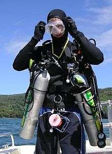 The front view of a standing diver ready for the water is shown. He is carrying a sling mounted aluminium cylinder on each side, clipped to a chest D-ring and a hip D-ring.