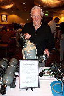 Eldred is holding a grey scuba cylinder and regulator with a sign titled "Australia's first scuba? The Porpoise scuba c 1953".