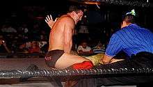 A dark-haired young man in a wrestling ring with black ropes. He is wearing black wrestling tights, with the name DiBiase written on them in red letters, and red kneepads with black wrestling boots. A referee wearing a blue shirt with thin black vertical stripes is in the corner.