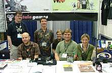 Five men and a woman in a convention booth for Telltale Games. A variety of Sam & Max merchandise is on the table in front of them.