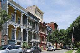 Rowhouses with arts-and-crafts styled porches (on both first and second floors) sit on a street across from a park.