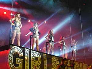 Five young women in bright leotards standing atop a sign reading "Girls Aloud"