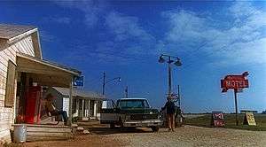 A flat, rural setting under a vast sky. On the left, a man sits on the porch of a small, old wooden building. In front of him, another man stands beside a pickup truck parked next to a gas station pump. On the right, a tall red sign reads "Mariposa Motel".