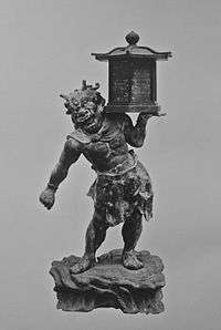  Tentōki. Front view of a stocky statue with a demon face. He is carrying a lantern on is left shoulder supported by his left hand. Black and white photograph.