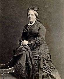 A photographic portrait of a woman with graying hair dressed in an elaborate dark mid-Victorian period dress and leaning against the back of an upholstered chair