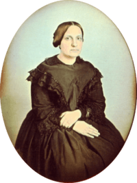 Photographic portrait of a woman seated and wearing a dark dress trimmed in dark lace, with her hair pulled back into a bun and no jewelry except for a simple ring on her left hand