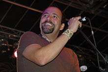 A man with a goatee and a T-shirt; he is smiling and his hand on a microphone stand