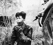 A scarred and bloodied man of Oriental appearance kneels on the ground. Beside him stands a man with an FN FAL battle rifle. A bayonet is fixed to the end of the rifle.