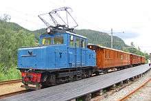 A small blue locomotive hauling three wooden passenger cars parked at a wooden platform