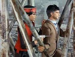 An agent dressed in a scarlet uniform and cap holds a suited civilian man hostage, positioning himself behind the other man so as to shield himself while pointing a gun at an unseen, off-screen enemy. The agent and hostage are standing on a metal structure of girders and supports, which is indicated to be high above ground level by the scale of the streets and buildings below them.