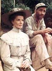 Hepburn is dressed in early-20th-century clothes, looking prim and proper. Behind her is Humphrey Bogart, also dressed as his character from The African Queen.