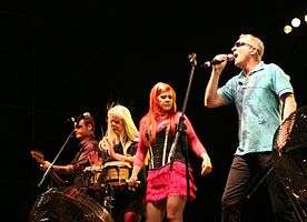 A group of musicians perform on a lighted stage. From left to right: a short-haired man plays bass guitar, a blond-haired woman plays the bongos, an red-haired woman holds a microphone, and a man wearing sunglasses and a blue shirt sings into a microphone.