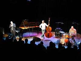 Stage with three musicians in their thirties. Left to right, man in a suit next to a Steinway grand piano, man in jeans holding a bass fiddle, man in jeans playing drums. Heads of audience visible in front.
