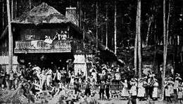 A theatre production, showing a crowd of villagers celebrating outside an inn