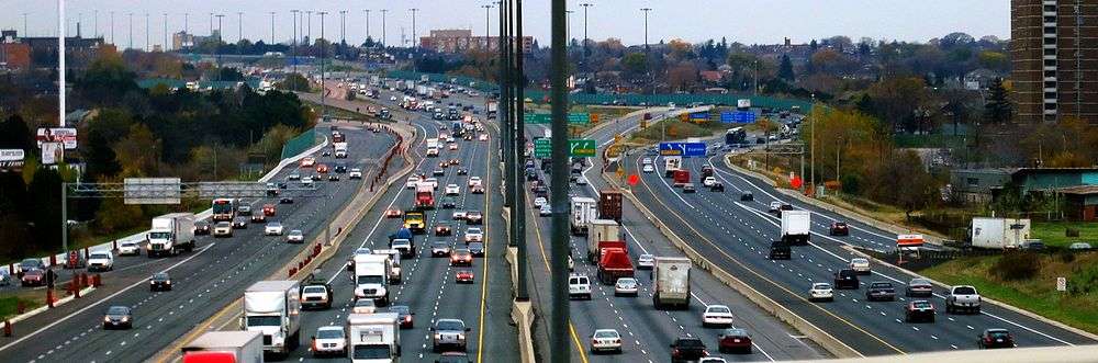 A wide angle image of Highway 401 completely occupied by cars. A city, Toronto, surrounds the freeway, but is walled off from it by green noise barriers. Each direction on the freeway is divided into two sets of lanes, referred to as a collector-express system.