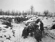In the foreground, a group of soldiers huddle in a group in a snow-covered trench. Another group stand in the background.