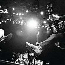 A black-and-white, worms-eye-view photo of two string players jumping in the air in front of a drum kit