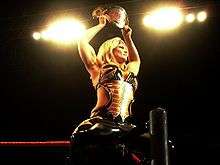 A blond woman sits on the turnbuckle of a wrestling ring, while facing away from the ring, with her legs on either side of the ring post. She is wearing a black and gold catsuit with the sides of the torso removed to bare part of her midriff, and is holding a wrestling championship in the air above her head.