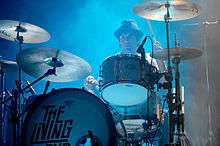 Strachan is mostly obscured by his drums. He wears a dark hat and stares ahead. His left hand has his drumstick high, while the right drumstick is low and over a cymbal.