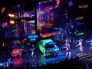 Bird's eye view of a stage, showing large scaffoldings, neon signs and a green car lying in the middle.