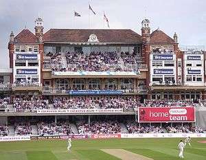 A three tiered Victorian red-brick building with flags flying on the roof and a clock at the top. There are large numbers of spectators watching a cricket match.