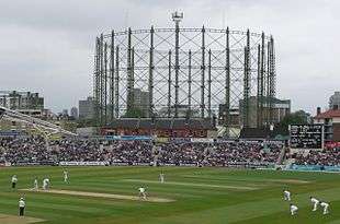 A cricket match in progress; gasholders are visible in the background
