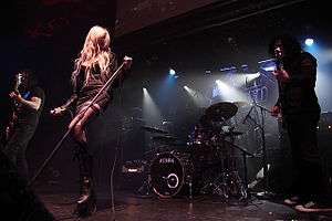 A tall woman with long blonde hair wearing a black dress and two men also wearing black performs in front of an audience.