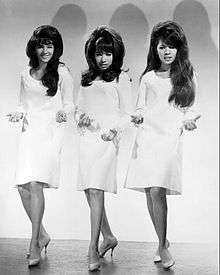 A black-and-white photograph of three women looking towards a camera. They all have large black hair and all wear short white dresses and heels.