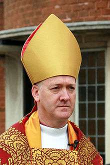 A solemn-looking middle-aged white man (head and shoulders shown), in bishop's robes – a gold mitre and red cope with gold highlights, standing in front of the windows of a red brick building.