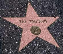 A coral-pink terrazzo five-point star inlaid into a charcoal-colored terrazzo background. In the upper portion of the pink star field, the text "The Simpsons" is inscripted. Below the inscription, in the lower half of the star field, there is a round emblem with an image of a television in it.